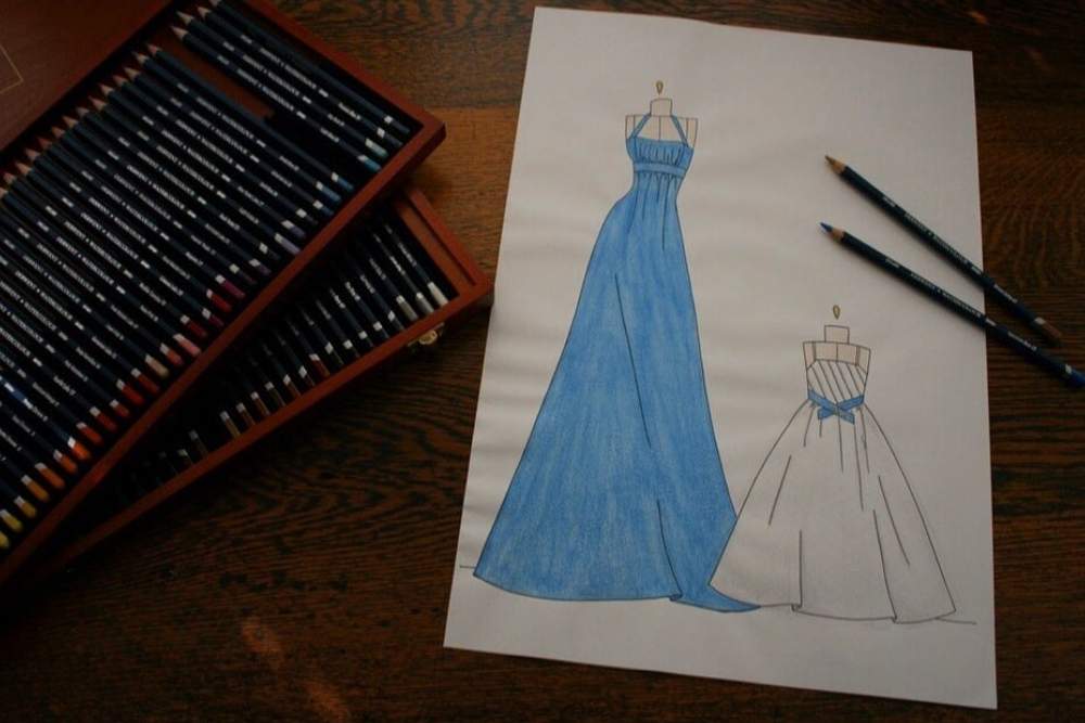 Hand drawn fashion illustrations of a blue and white dress on a wooden desk next to a box of pencils-